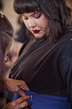 A person in a black outfit is having their blue garment adjusted by another individual. The focus is on the process of fastening or zipping up the clothing, with attention to detail in the clothing and the hands assisting.