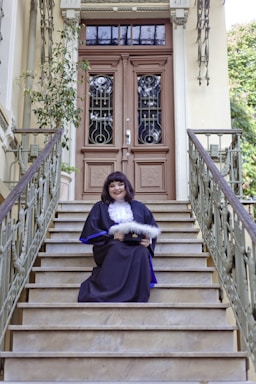 A person in formal academic attire sits on the steps of a grand entrance with ornate double doors. The railing and surrounding architecture feature intricate designs. A potted plant decorates the side of the entrance.