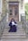 A person in formal academic attire sits on the steps of a grand entrance with ornate double doors. The railing and surrounding architecture feature intricate designs. A potted plant decorates the side of the entrance.