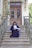 A person in formal academic attire sits on the steps of a grand entrance with ornate double doors. The railing and surrounding architecture feature intricate designs. A potted plant decorates the side of the entrance.