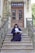 A person in formal academic attire sits on the steps of a grand entrance with ornate double doors. The railing and surrounding architecture feature intricate designs. A potted plant decorates the side of the entrance.