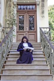 A person in formal academic attire sits on the steps of a grand entrance with ornate double doors. The railing and surrounding architecture feature intricate designs. A potted plant decorates the side of the entrance.