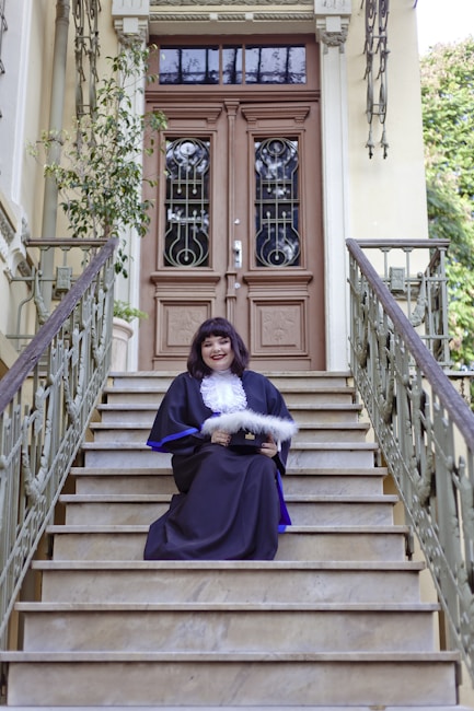 A person in formal academic attire sits on the steps of a grand entrance with ornate double doors. The railing and surrounding architecture feature intricate designs. A potted plant decorates the side of the entrance.