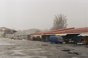 An overcast outdoor market area with a series of single-story stalls covered by red and yellow awnings. There is a blue van parked in front of one of the stalls, and a few vendors setting up or attending their merchandise. The ground is wet with puddles. In the background, there is a large tree without leaves and some industrial buildings or warehouses.