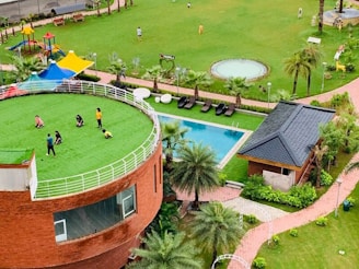 A lush green open space features a circular building with a grassy rooftop where several people are gathered, likely children and adults. The area is complemented by a playground with colorful structures, a swimming pool, and landscaped gardens with palm trees. Nearby is a small brick building with a dark roof.