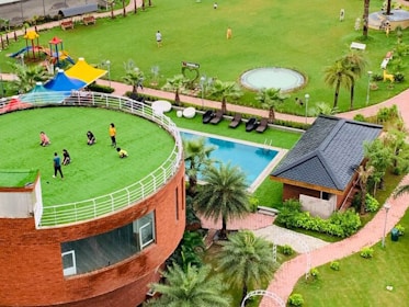 A lush green open space features a circular building with a grassy rooftop where several people are gathered, likely children and adults. The area is complemented by a playground with colorful structures, a swimming pool, and landscaped gardens with palm trees. Nearby is a small brick building with a dark roof.