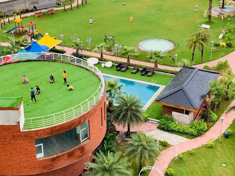 A lush green open space features a circular building with a grassy rooftop where several people are gathered, likely children and adults. The area is complemented by a playground with colorful structures, a swimming pool, and landscaped gardens with palm trees. Nearby is a small brick building with a dark roof.