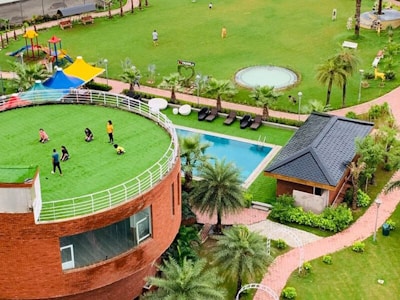A lush green open space features a circular building with a grassy rooftop where several people are gathered, likely children and adults. The area is complemented by a playground with colorful structures, a swimming pool, and landscaped gardens with palm trees. Nearby is a small brick building with a dark roof.