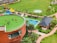 A lush green open space features a circular building with a grassy rooftop where several people are gathered, likely children and adults. The area is complemented by a playground with colorful structures, a swimming pool, and landscaped gardens with palm trees. Nearby is a small brick building with a dark roof.