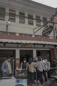 A small group of people gathers outside a traditional coffee shop with a vintage sign that reads 'Warung Kopi Purnama.' The customers, mostly young adults, are wearing casual clothing and face masks. One person is operating a cart with bread or pastries in a display case. The building has colorful louvered windows above the entrance, adding a quaint charm.