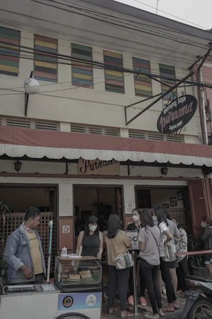 A small group of people gathers outside a traditional coffee shop with a vintage sign that reads 'Warung Kopi Purnama.' The customers, mostly young adults, are wearing casual clothing and face masks. One person is operating a cart with bread or pastries in a display case. The building has colorful louvered windows above the entrance, adding a quaint charm.