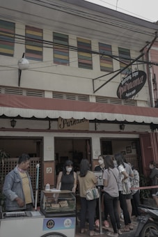 A small group of people gathers outside a traditional coffee shop with a vintage sign that reads 'Warung Kopi Purnama.' The customers, mostly young adults, are wearing casual clothing and face masks. One person is operating a cart with bread or pastries in a display case. The building has colorful louvered windows above the entrance, adding a quaint charm.
