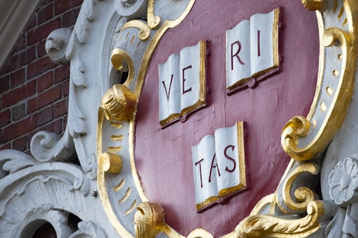A sculpted emblem with the Latin word 'VERITAS' displayed on three open books. The emblem features a shield shape with ornate gold and white decoration, set against a brick background.