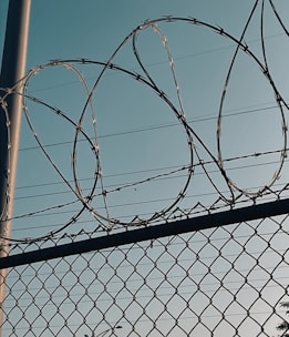 Barbed wire is coiled above a metal chain-link fence, with a clear sky in the background. The image conveys a sense of security and restriction.