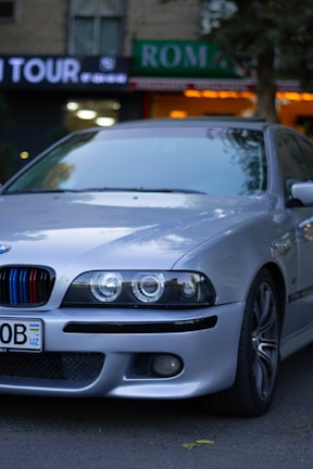 A silver BMW car is parked on the street. The vehicle has a custom front grille with red and blue stripes, and sleek, modern headlights. In the background, there are various storefronts, including one with a green 'ROMA' sign and another sign that reads 'TOUR'. The surroundings include some trees and city elements, like building façades.
