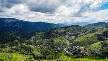 A breathtaking landscape of terraced rice fields sprawling across lush green hills, dotted with small buildings and interspersed with dense forests under a partially cloudy sky. Mountains in the distance frame the scene, adding depth to the panoramic view.