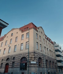 A historic multi-story building with a beige brick facade and maroon roof is set against a clear blue sky. The architecture includes arched windows and decorative stonework along the ground floor. The building is adjacent to a more modern structure with white balconies.