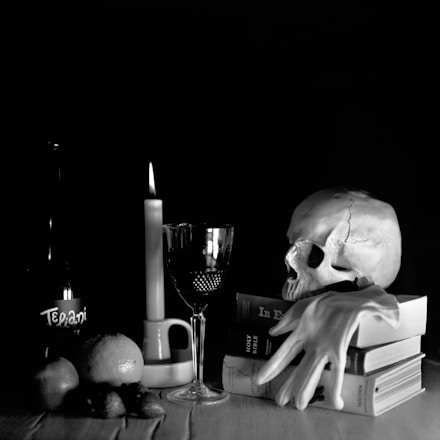 A still life arrangement features a human skull resting on a stack of books, with an elegant wine glass and a lit candle nearby. A bottle with the label partially visible stands beside a bowl containing fruits, including an orange and some smaller fruits. Draped over the books is a white glove, contributing to a mysterious undertone.