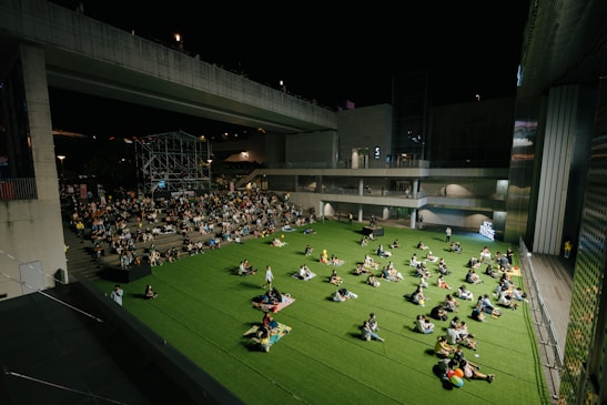 A nighttime outdoor event taking place in an urban setting. People are seated on a large area of artificial grass, comfortably spaced apart. Some sit in groups, while others are alone, creating a relaxed and communal atmosphere. A stage is visible in the background, surrounded by a crowd seated on steps. The scene is framed by modern architecture featuring large, concrete structures and ambient lighting.