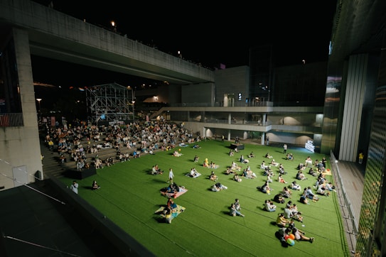 A nighttime outdoor event taking place in an urban setting. People are seated on a large area of artificial grass, comfortably spaced apart. Some sit in groups, while others are alone, creating a relaxed and communal atmosphere. A stage is visible in the background, surrounded by a crowd seated on steps. The scene is framed by modern architecture featuring large, concrete structures and ambient lighting.