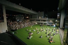A nighttime outdoor event taking place in an urban setting. People are seated on a large area of artificial grass, comfortably spaced apart. Some sit in groups, while others are alone, creating a relaxed and communal atmosphere. A stage is visible in the background, surrounded by a crowd seated on steps. The scene is framed by modern architecture featuring large, concrete structures and ambient lighting.