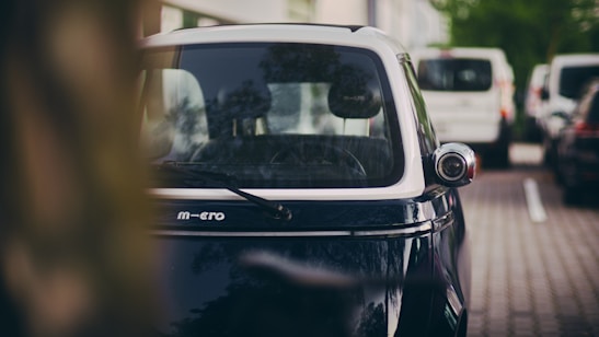 A small, compact electric vehicle is parked on the side of a street, partially obscured by a tree trunk in the foreground. The vehicle has a sleek, modern design with a glossy finish, and its windshield is reflective. In the background, several larger vehicles are parked on the street, with greenery and buildings visible, suggesting an urban environment.