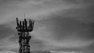 A black and white image of a communication tower with antennas and dishes against a cloudy sky.