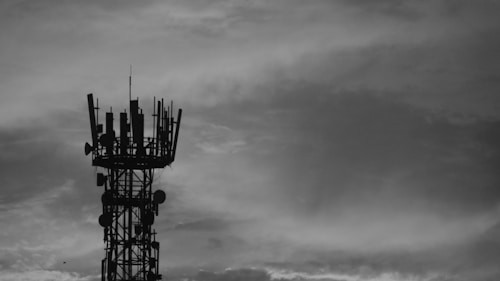 A black and white image of a communication tower with antennas and dishes against a cloudy sky.