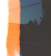 A photograph that appears to have a light leak effect, creating a strong division of color within the frame. On the left side, there is a bright orange and white area, while the right side has a darker section with silhouetted palm trees and electrical wires against a deep blue sky. The overall composition evokes a sense of abstraction and nostalgia, possibly indicating a film photography effect.