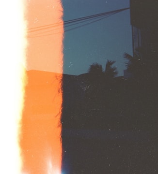A photograph that appears to have a light leak effect, creating a strong division of color within the frame. On the left side, there is a bright orange and white area, while the right side has a darker section with silhouetted palm trees and electrical wires against a deep blue sky. The overall composition evokes a sense of abstraction and nostalgia, possibly indicating a film photography effect.