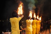A group of people wearing yellow garments hold large, multi-tiered brass lamps with flames, standing in a line during a nighttime ritual or ceremony. The scene is set by a river, and a small crowd of spectators can be seen near the edge. The background is dark with scattered lights visible in the distance.