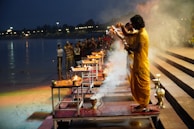 A group of people are performing a religious ceremony by a riverbank. Individuals dressed in yellow robes hold ritual items in the air as part of the observance. Smoke from incense creates a mystical atmosphere, while onlookers watch the event. The scene is set at dusk with lights reflecting on the water.