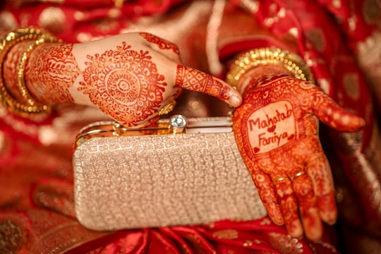 A close-up view of hands adorned with intricate henna designs. One hand holds a golden clutch purse, while the other displays the names 'Mahtab' and 'Fariya' within a decorative henna pattern. The background includes red fabric with gold accents, enhancing the traditional aesthetic.