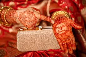 A close-up view of hands adorned with intricate henna designs. One hand holds a golden clutch purse, while the other displays the names 'Mahtab' and 'Fariya' within a decorative henna pattern. The background includes red fabric with gold accents, enhancing the traditional aesthetic.