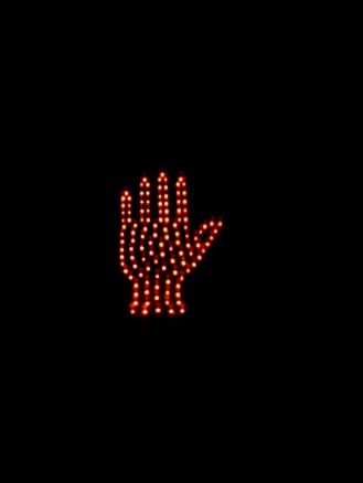 A digital stop sign in the shape of a hand is made up of numerous red LED lights against a dark background. The hand symbol is illuminated, clearly signaling to stop.