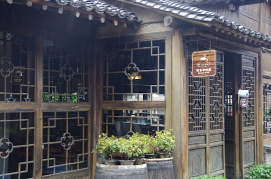 A traditional wooden building with intricate lattice window designs and a tiled roof, showcasing a cozy corner cafe. Potted plants are placed in rustic barrels in front of the entrance, and a wooden sign with a steaming cup symbol hangs above the door.