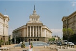 A grand neoclassical building with a prominent central spire dominates the image. The building is symmetrical with architectural details typical of government or institutional structures. A glass domed structure is in the foreground, surrounded by a pedestrian area with planters. Flanking the central building are similarly styled structures, and several vehicles are visible on the street, suggesting an urban setting.