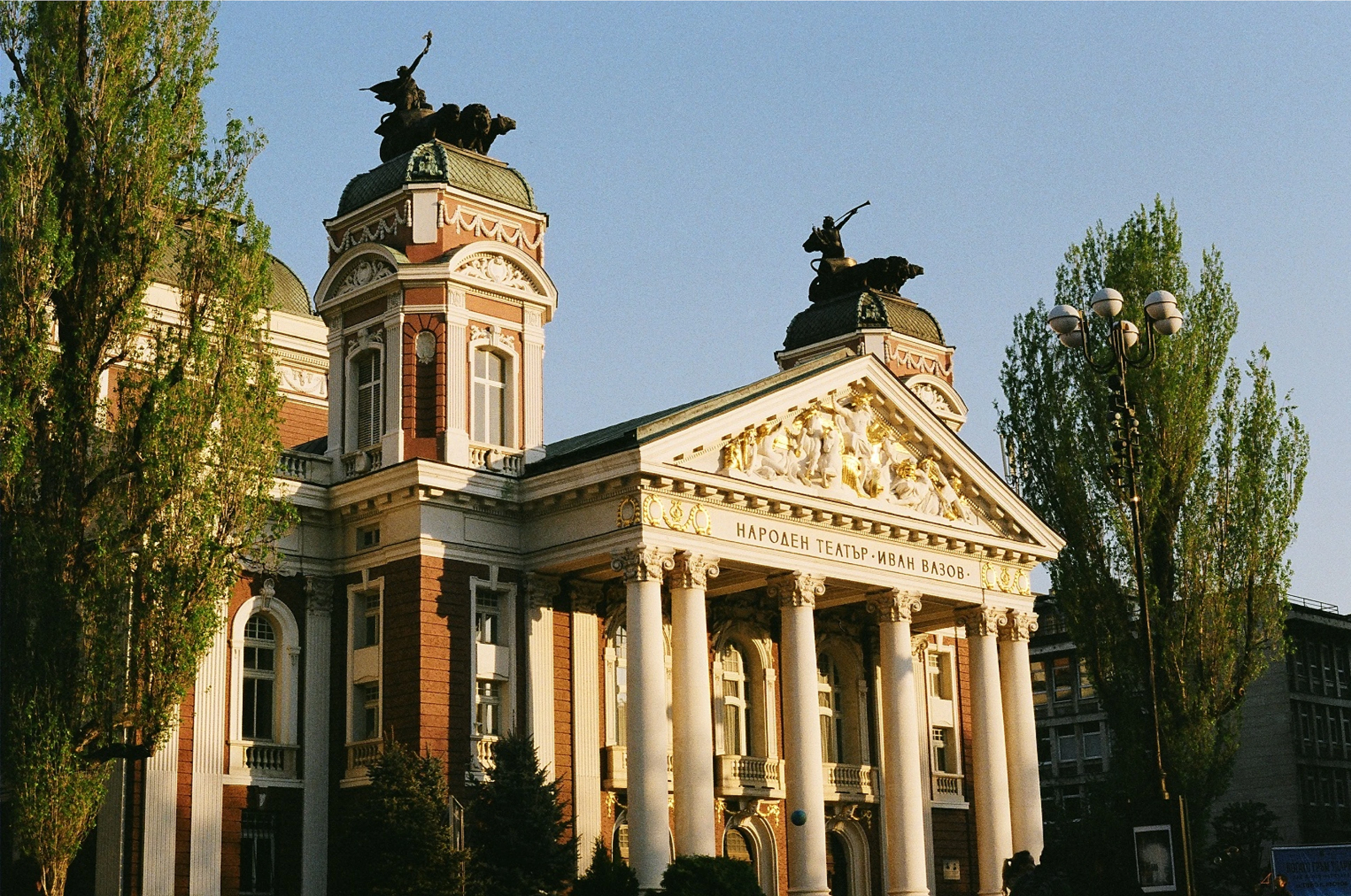 The columned portico and bronze quadrigas of the Ivan Vazov National Theatre in Sofia's City Garden, its gilded pediment catching low evening sun between tall poplars.