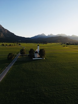 A lone, small white church with a dark roof surrounded by a few trees is situated in a vast green field. The setting is in the countryside with a straight road leading towards the church. Mountains are visible in the background under a clear blue sky.