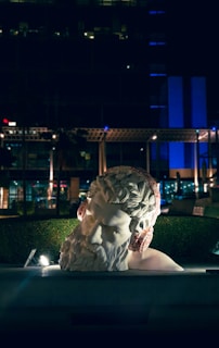 A large classical sculpture of a bearded male head with red headphones is placed in an outdoor setting, surrounded by hedges and modern structures. The image is taken at night, with artificial lighting highlighting the sculpture against a dark background.