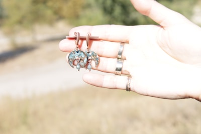 A pair of crescent-shaped earrings with turquoise accents is held in a hand against a blurred outdoor background. The earrings have intricate designs with small dangling elements. Several rings adorn the fingers of the hand.