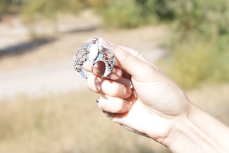 A hand is holding an ornate silver ring with intricate designs against a blurred outdoor background.