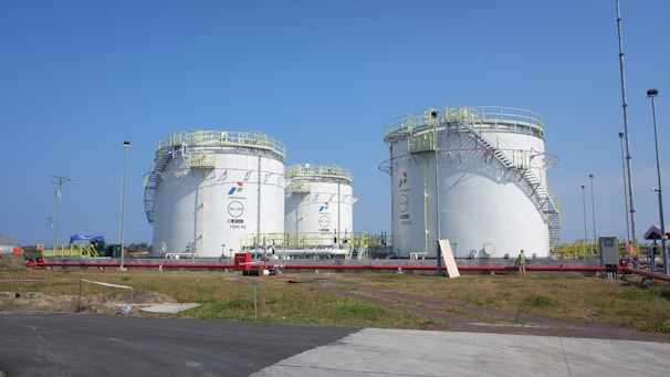 Several large, industrial storage tanks are situated on a grassy area, equipped with safety railings and exterior staircases. These tanks are part of a storage facility under a clear blue sky.
