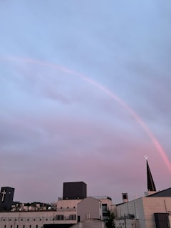 A cityscape featuring several modern buildings with a prominent church steeple displaying a cross. Above, a pale rainbow arches across a sky filled with soft pastel hues of pink and blue-grey clouds, creating a serene atmosphere.