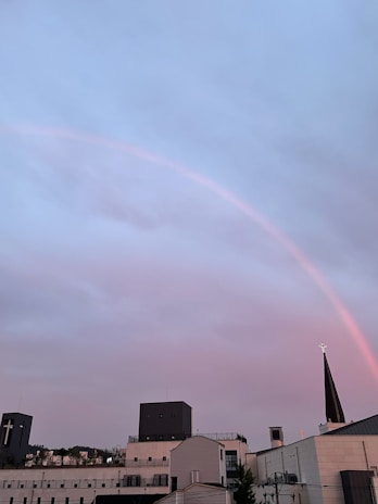 A cityscape featuring several modern buildings with a prominent church steeple displaying a cross. Above, a pale rainbow arches across a sky filled with soft pastel hues of pink and blue-grey clouds, creating a serene atmosphere.
