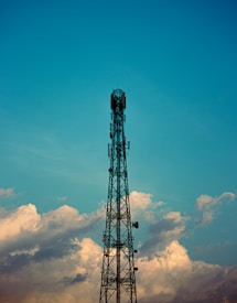 A tall communication tower stands against a backdrop of a blue sky with scattered clouds. The structure is made of metal and rises prominently from the bottom to the top center of the image.
