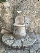 A rustic water fountain with a sign above it reading 'eau potable', set against a textured stone wall. The fountain is made of stone with small bird carvings and sits atop a circular stone platform.