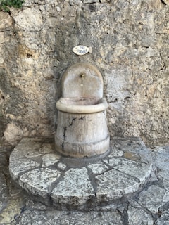 A rustic water fountain with a sign above it reading 'eau potable', set against a textured stone wall. The fountain is made of stone with small bird carvings and sits atop a circular stone platform.