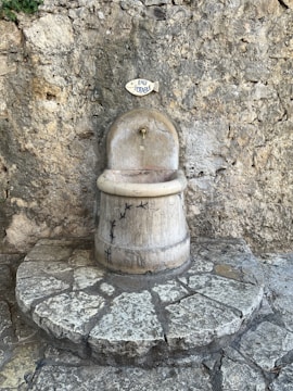 A rustic water fountain with a sign above it reading 'eau potable', set against a textured stone wall. The fountain is made of stone with small bird carvings and sits atop a circular stone platform.