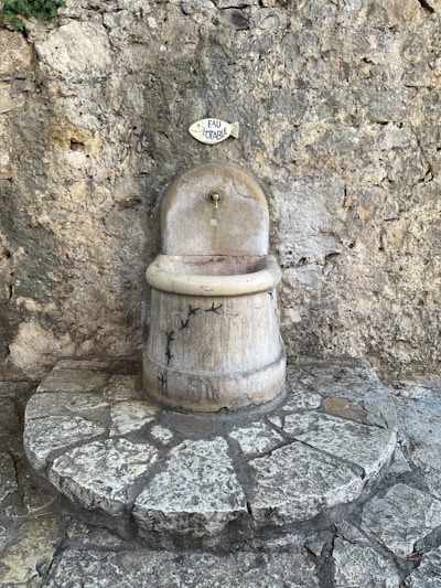 A rustic water fountain with a sign above it reading 'eau potable', set against a textured stone wall. The fountain is made of stone with small bird carvings and sits atop a circular stone platform.